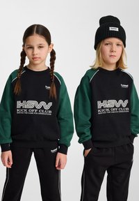 Svart och grön sweatshirt med texten "HSW KICK OFF CLUB", ribbstickade ärmar och avslappnad passform, tillsammans med svarta byxor och en ribbad svart mössa.