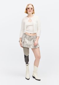 Femme aux cheveux blonds courts portant des lunettes de soleil blanches, un cardigan court blanc, une jupe à motifs et une jambe prothétique avec des bottes blanches.