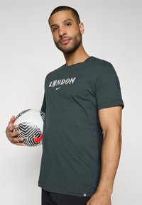 Tumši zaļš T-krekls ar uzrakstu "LONDON" un Nike logo priekšpusē. Modelis tur melnbaltu teksturētu futbola bumbu ar sarkanu akcentu.