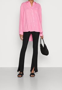 Light pink long-sleeve shirt with front buttons and a pocket, paired with black fitted pants featuring side slits, and black woven handbag.