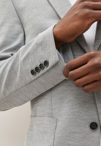 Blazer gris clair avec finition texturée. Comprend quatre boutons noirs sur le poignet et une poche à l'avant, avec une doublure rayée visible.