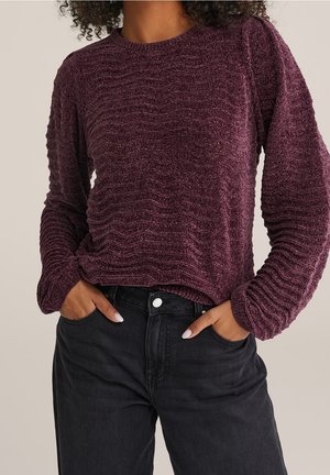 Personne portant un pull violet texturé à manches longues avec des motifs ondulés subtils, associé à un jean noir taille haute, les mains dans les poches.
