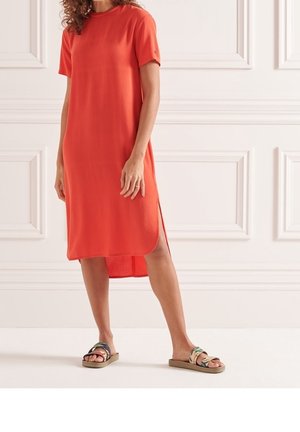 Femme portant une robe orange mi-longue à manches courtes avec une fente sur le côté et des sandales plates aux brides multicolores.