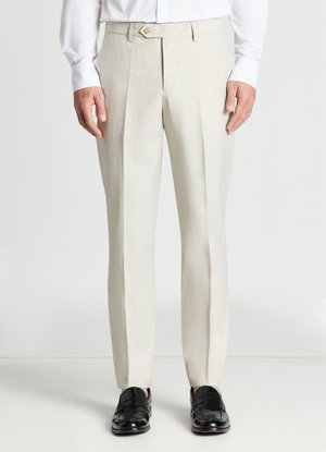 Homme portant un pantalon beige sur mesure avec des plis à l'avant, une chemise blanche et des mocassins en cuir noir, debout devant un fond uni.