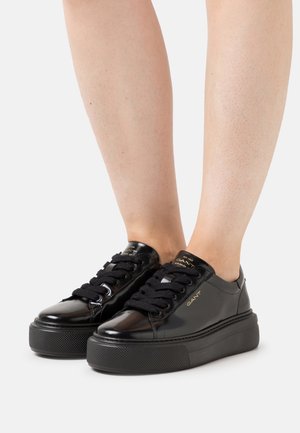 Sneakers piattaforma nere lucide con lacci neri indossate su gambe nude contro uno sfondo bianco, con il logo GANT in oro.
