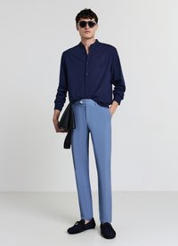 Chemise bleu marine à manches longues, pantalon sur mesure bleu clair, lunettes de soleil noires et pochette noire. Les chaussures sont bleu marine avec une finition texturée.