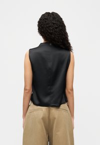 Μαύρο αμάνικο τοπ από λείο ύφασμα με ψηλό λαιμόκοψη και ελαφρώς κυρτή γραμμή στο τελείωμα, συνδυασμένο με μπεζ παντελόνι. Χωρίς ορατά λογότυπα.