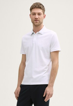 Witte polo met een klassieke kraag, korte mouwen en een subtiel textuurpatroon. Gedragen met donkere broeken. Minimale branding zichtbaar.
