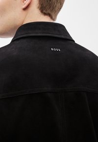 Melnās zamšādas jaka ar strukturētu apkakli, uz muguras ar baltu, nepamanāmu logotipu "BOSS". Gluda tekstūra un tīras šuvju detaļas.