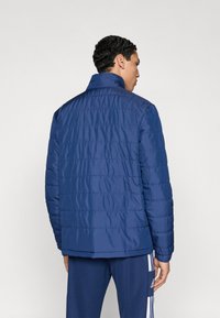 Μπλε σκούρο μπουφάν πάππουερ με ψηλό γιακά, καπιτονέ σχέδιο και ελαστικά μανίκια, φορεμένο πάνω από μπλε αθλητικό παντελόνι. Ομαλή υφή υφάσματος.