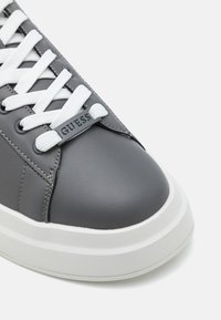 Baskets en cuir gris avec des lacets blancs, un bout arrondi et une épaisse semelle blanche. Présente un onglet de marque sur la languette avec le texte "GUESS".