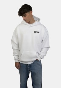Felpa bianca con cappuccio e scritta nera "CRYSTAL". Presenta una tasca frontale e una vestibilità ampia, abbinata a jeans azzurri chiari. Materiale morbido in cotone.