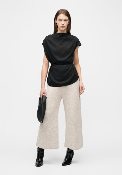 Γυναίκα που φοράει χαλαρό μαύρο αμάνικο τοπ, μπεζ πλεκτό παντελόνι με φαρδύ πόδι, μαύρα μποτάκια μέχρι τον αστράγαλο και κρατάει μια μαύρη τσάντα χειρός.