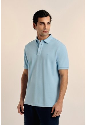 Hombre con una camiseta polo de manga corta azul claro, de pie contra un fondo liso y claro, mirando hacia un lado.