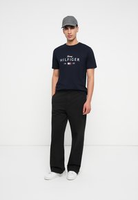 Námořnické bavlněné tričko s logem "Tommy HILFIGER", doplněné černými kalhotami a šedou čepicí. Boty jsou bílé, s minimalistickým designem.