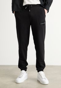Zwarte sweatpants met elastische boorden, een trekkoord in de taille en geborduurd logo. Gepaard met witte sneakers op een neutrale vloer.