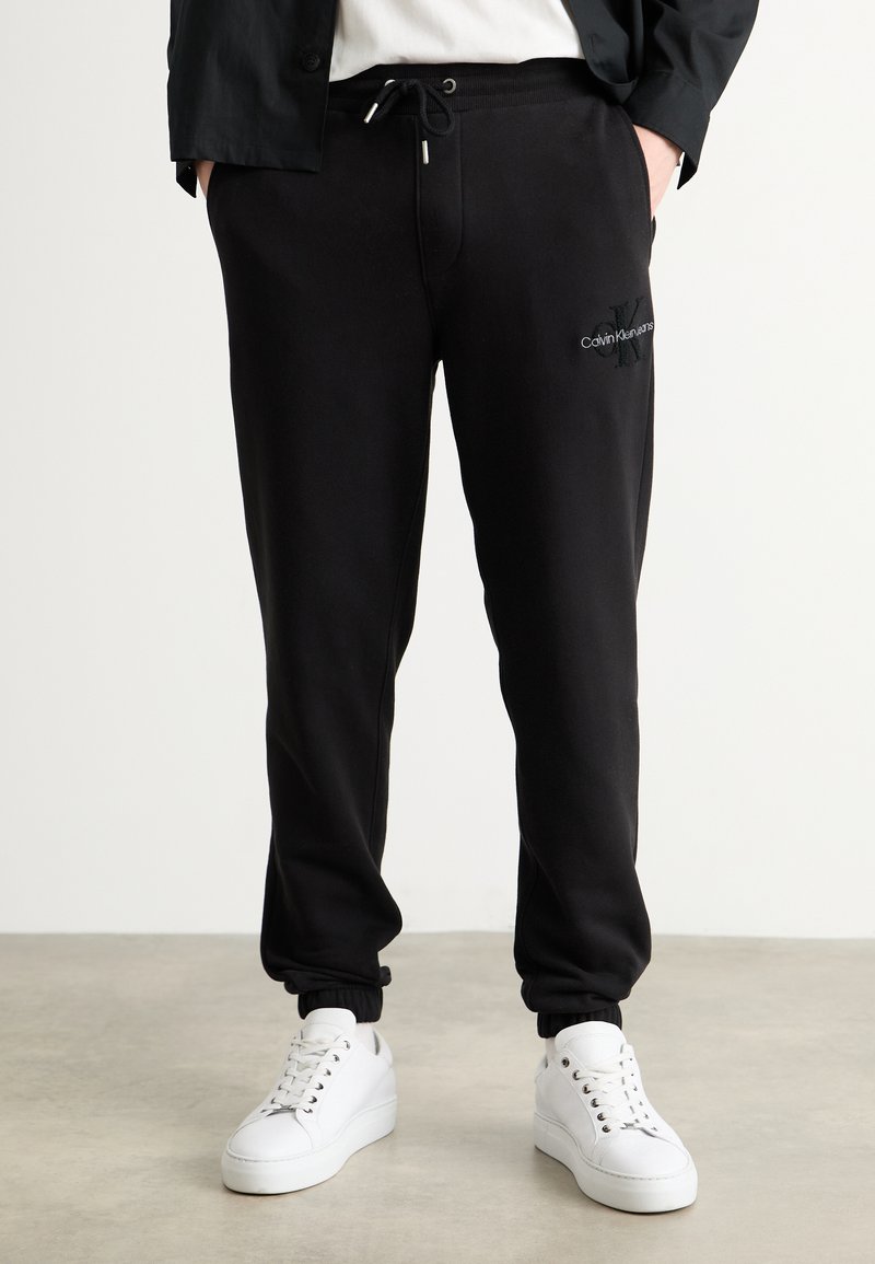 Zwarte sweatpants met elastische boorden, een trekkoord in de taille en geborduurd logo. Gepaard met witte sneakers op een neutrale vloer.