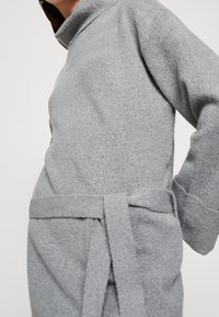 Robe-pull gris clair avec un col montant, des manches longues et une ceinture nouée à la taille. Tissu texturé avec une finition douce et brossée.