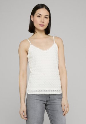 Kvinde med glat mørkt hår iført en hvid tekstureret ærmeløs top med tynde stropper og grå jeans mod en ensfarvet grå baggrund.