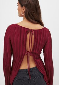 Bordeaux langærmet bluse med lodrette plisseringer, åben rygdesign bundet om halsen og asymmetrisk kant. Glat stof med en subtil glans.