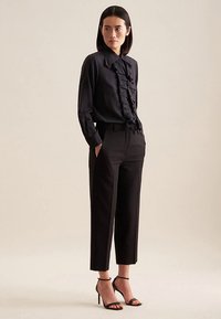 Camicia nera con dettagli a volant, abbinata a pantaloni neri su misura. Modella con tacchi neri con cinturini. Tessuto liscio, design aderente.