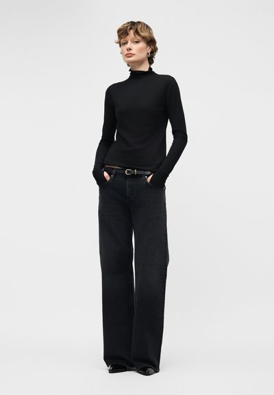 Γυναίκα όρθια με χέρια στις τσέπες, φοράει μαύρο μακρυμάνικο πουλόβερ με ζιβάγκο και μαύρα τζιν παντελόνια φαρδιάς γραμμής, μπροστά σε απλό φόντο.