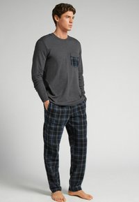 Camiseta gris de manga larga con un bolsillo que presenta un diseño de cuadros azules, combinada con pantalones de pijama de cuadros azules y negros. No se llevan zapatos.