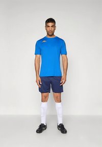 Μπλε αθλητική μπλούζα με κοντά μανίκια, σε συνδυασμό με σκούρο μπλε σορτς, λευκές κάλτσες ως το γόνατο και μαύρα ποδοσφαιρικά παπούτσια, με λογότυπο στο στήθος.