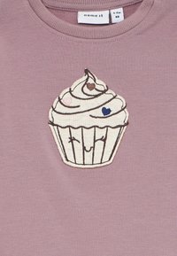 Maglietta lilla per bambini con ricamo di una torta a forma di cupcake sorridente, decorata con piccoli cuori rosa, marroni e blu.