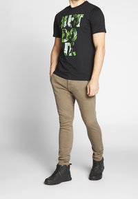 Czarna koszulka z zielonym napisem "JUST DO IT" ozdobionym wzorem liści, zestawiona z jasnobeżowymi, dopasowanymi spodniami i czarnymi sznurowanymi trampkami.