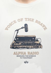 Εικονογράφηση με ρετρό ραδιόφωνο και μικρόφωνο με τα κείμενα «Φωνή των Γενναίων», «Alpha Radio» και «Φέρνοντας την αποστολή στον δρόμο».