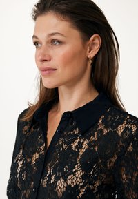 Zwarte kanten blouse met een effen zwarte kraag en knopen. Bloemenpatroon is zichtbaar door de kant. Model toont een profielzicht.