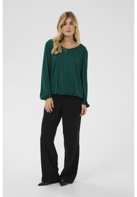 Dunkelgrünes Langarmoberteil mit rundem Ausschnitt, kombiniert mit schwarzen, weit geschnittenen Hosen und spitzen schwarzen Schuhen, vor neutralem Hintergrund.