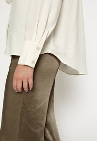 Kreemjateksiline pluus nupuvarrukatega, millele on sobitatud siidised oliivrohelised püksid, näidates siledat kangast ja mugavat lõiget.