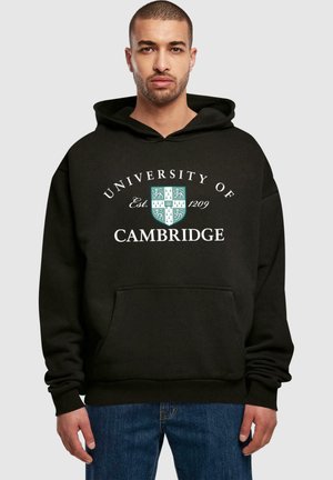 Black hoodie with a front pocket, featuring white text reading "University of Cambridge" and a green shield design. Loose fit.