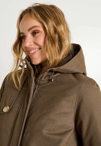 Chaqueta exterior marrón con capucha estructurada, cierre de cremallera y un gran detalle de botón. Tela texturizada con costuras visibles.