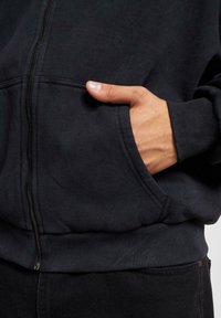 Main partiellement dans la poche avant d'un sweat à capuche noir zippé porté avec un pantalon noir.