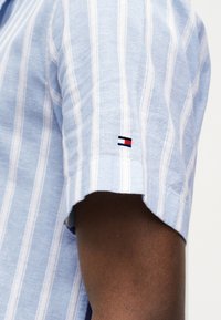 Hellblau-weiß gestreiftes Kurzarmhemd auf einem dunkelhäutigen Arm, mit einem kleinen, bestickten Logo in Rot, Weiß und Marineblau.