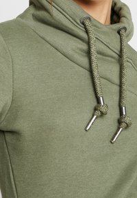 Primo piano di un tessuto di felpa verde con spesse stringhe di chiusura annodate vicino a punte metalliche argentate attorno all'alta colletto.