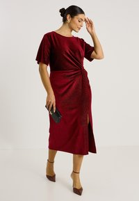 Robe en velours rouge profond avec manches courtes, détail de taille torsadée et fente latérale. Associée à une pochette noire et des escarpins marron à bouts pointus.