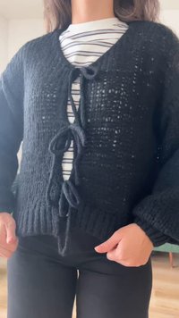 Persoon draagt een zwarte gebreide cardigan, gestrikt met drie strikken, over een wit-zwart gestreept shirt, staand binnen.