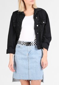 Giacca di jeans nera con bottoni frontali e maniche arrotolate, abbinata a una gonna di jeans azzurro chiaro con orlo sfibrato e una cintura a scacchi.