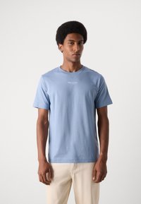 Tricou din bumbac de culoare albastru deschis, cu mâneci scurte și guler rotund. Prezentă un mic logo alb cu text pe piept. Textură netedă.