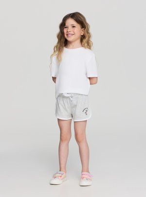 Ragazza giovane sorridente con capelli lunghi e ondulati, che indossa una t-shirt bianca semplice, pantaloncini grigio chiaro con logo di palma e sneakers pastello.