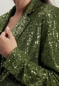 Groene paillettenblazer met een gestructureerde afwerking, sjaalkraag en opvallende glans. Close-up van de manchetten en hand op de revers.