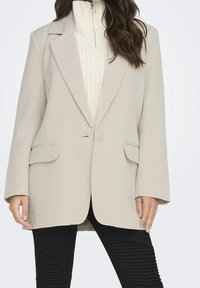 Blazer beige con un cierre de un solo botón, solapas en pico y dos bolsillos frontales, llevado sobre un suéter crema texturizado y pantalones negros de canalé.
