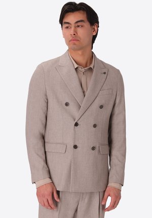 Uomo che indossa una giacca doppiopetto beige e pantaloni abbinati, guarda di lato contro uno sfondo chiaro e uniforme.