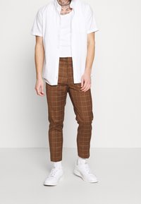 White short-sleeve shirt over a brown check-patterned pant, featuring a slim fit and ankle-length cut, paired with white sneakers.