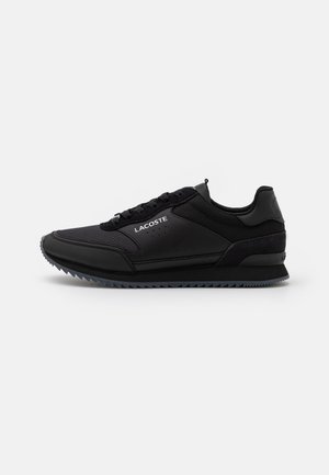 Baskets basses noires Lacoste avec panneaux en mesh et cuir, fermeture à lacets et semelle texturée, vues de côté sur fond blanc.