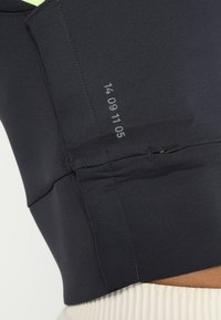 Czarny sportowy top o dopasowanym kroju, wykonany z teksturowanej tkaniny, z małą kieszonką na zamek. Zawiera szczegóły w postaci numerowego nadruku po boku.